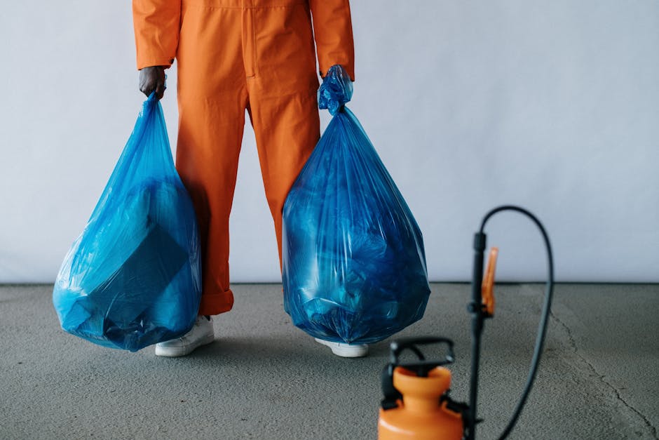 A person wearing orange work trousers and white sneakers is standing on a grey indoor floor, holding two large blue plastic rubbish bags filled with waste material, with one bag in each hand. The back