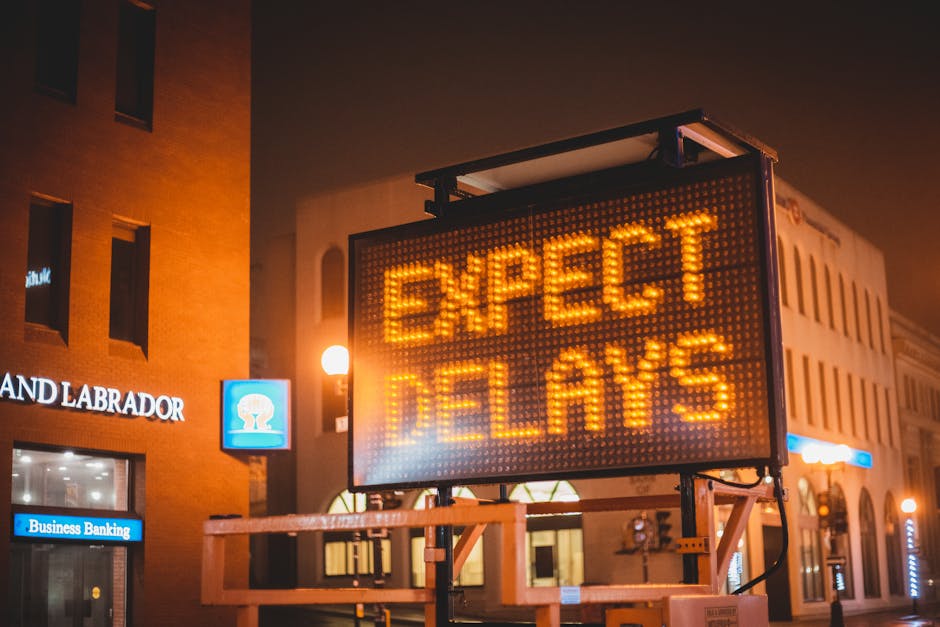 A large, illuminated electronic traffic sign displaying the message 'EXPECT DELAYS' in bright orange LED lights, positioned on a street corner at night. The sign is mounted on a metal frame and is sit
